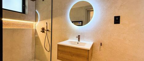 A modern bathroom with a glass-enclosed shower, a wooden vanity, and a round mirror with a light.