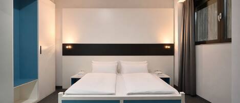 A modern hotel room with a double bed, a wardrobe, a bedside table, and a window with curtains.