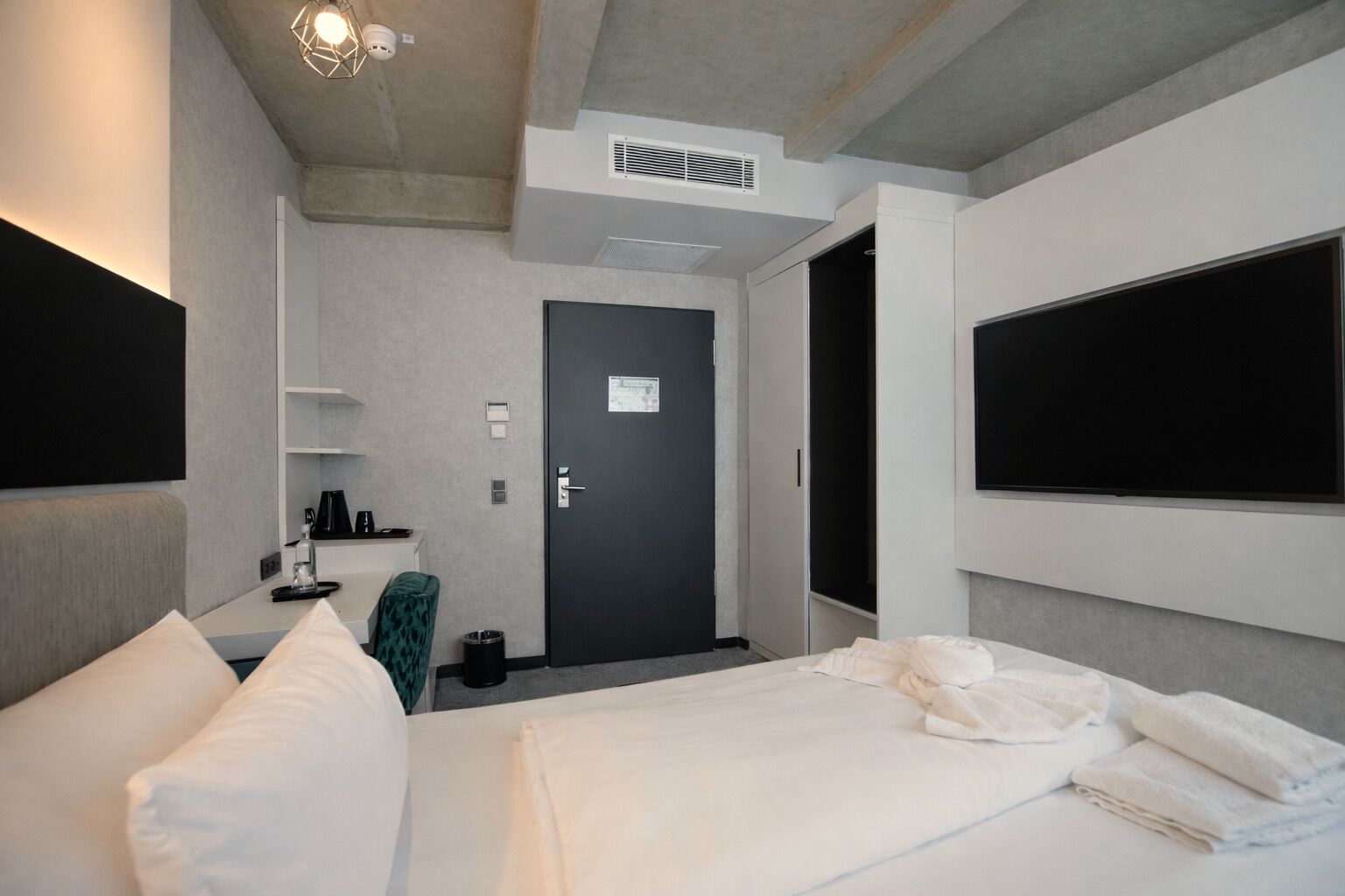 Ein modernes Hotelzimmer mit einem großen Bett, einem flachbildfernseher an der Wand, einem Schreibtisch mit Stuhl und einem Kleiderschrank.