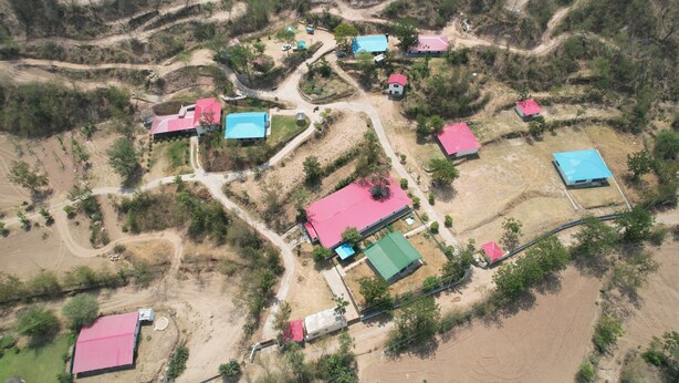 Vue aérienne d’une zone rurale avec plusieurs bâtiments, dont des maisons et une plus grande construction dotée d’un toit rouge, le tout entouré de champs en terrasses et d’une végétation clairsemée.