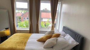 Una cama bien hecha con un edredón amarillo, almohadas blancas y un cabecero gris en una habitación con ventanas amplias y cortinas.