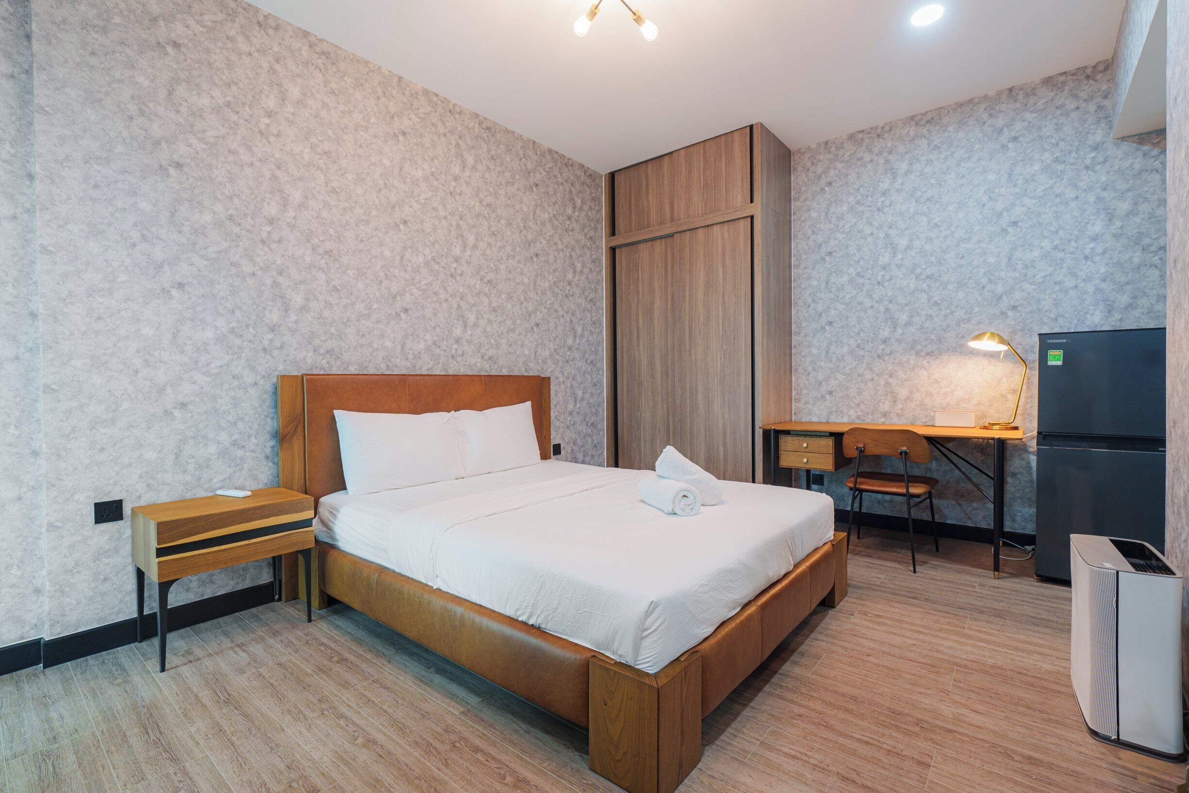 Ein Hotelzimmer mit einem hölzernen Bett, einem Schreibtisch mit Stuhl, einem Kühlschrank und einer Mülltonne.