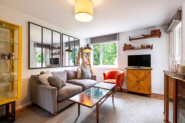 Sala de estar com sofá, mesa de centro e televisão. Estantes com objetos decorativos e uma janela com vista para o verde.