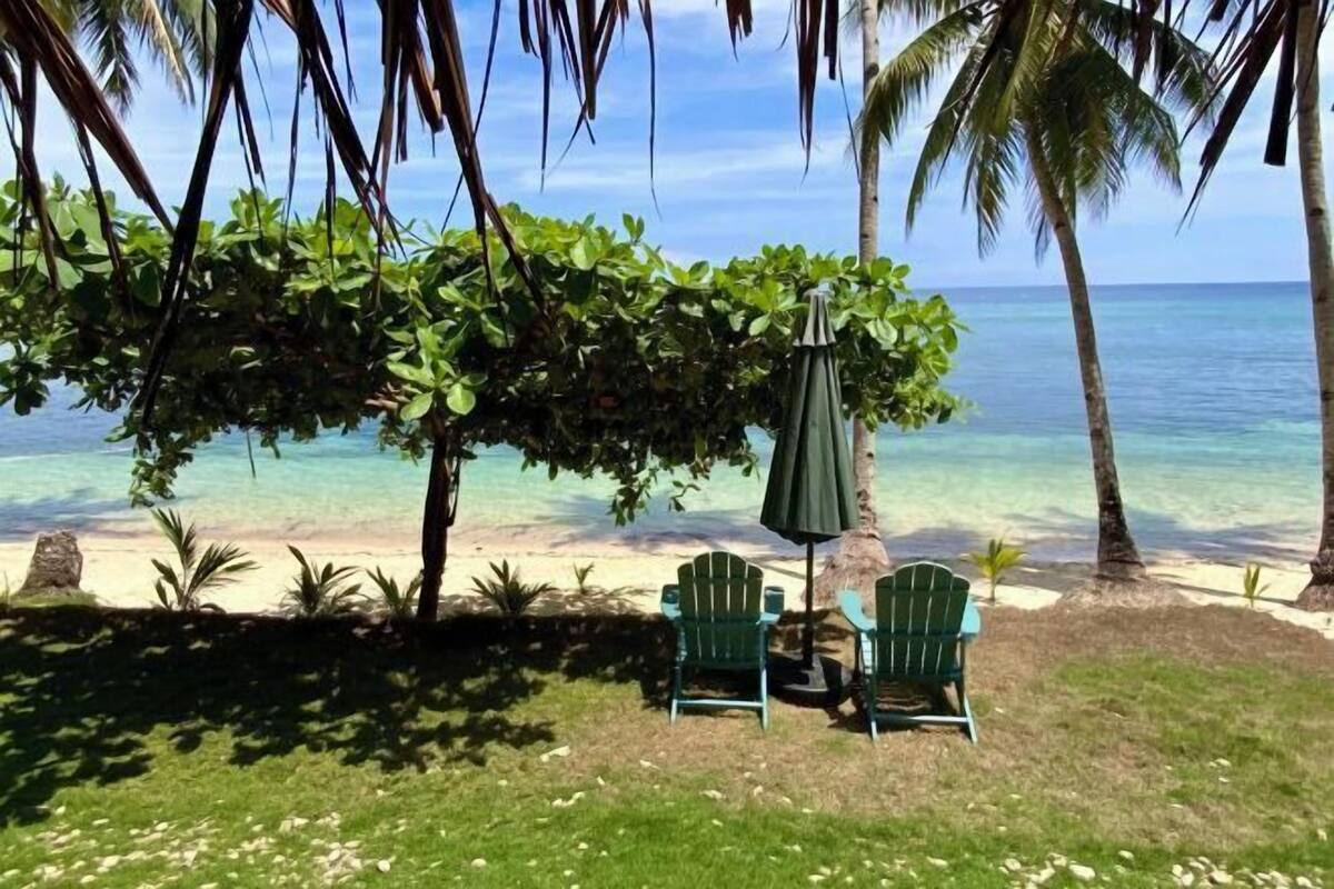 Un área de estar frente a la playa con sillas y una sombrilla, con vista al océano azul y transparente.