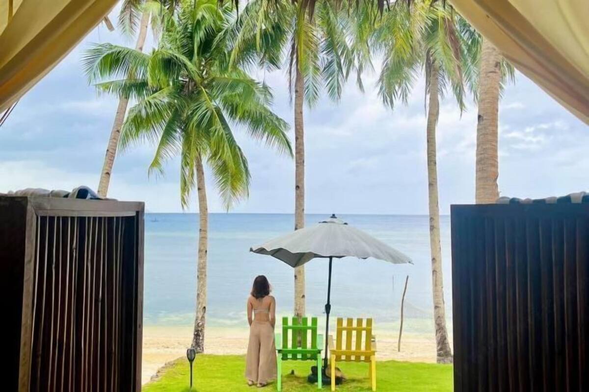 Vista de una playa con palmeras, una mujer bajo una sombrilla y sillas de colores.