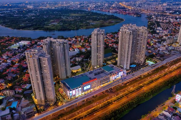 A high-rise building with multiple floors, a large commercial area with illuminated signage, and a river in the background. - Genius Saigon - Masteri Thao Dien District 2 (Ho Chi Minh City)