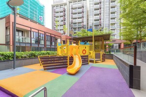 A playground with a yellow slide and a climbing structure, surrounded by a colorful play area and modern buildings in the background. - The Metropole - Horizon View Riverside  (Ho Chi Minh City)