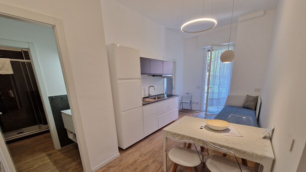 A compact kitchen with white cabinets, a dining area with two chairs, a small table, and a view of a bedroom with a bed and a window. - Apartment 4 (Milano)