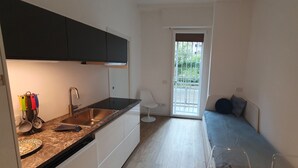 A compact kitchen with a sink, stove, and cabinets, a small white chair, and a sofa bed with a view to a balcony. - Apartment 4 (Milano)
