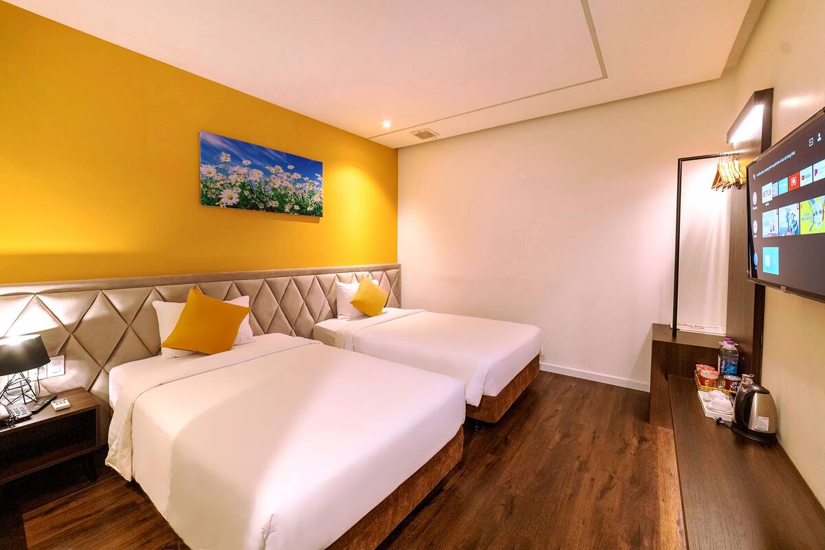 A hotel room with two beds, a painting on the wall, a TV on the wall, and a wooden floor.