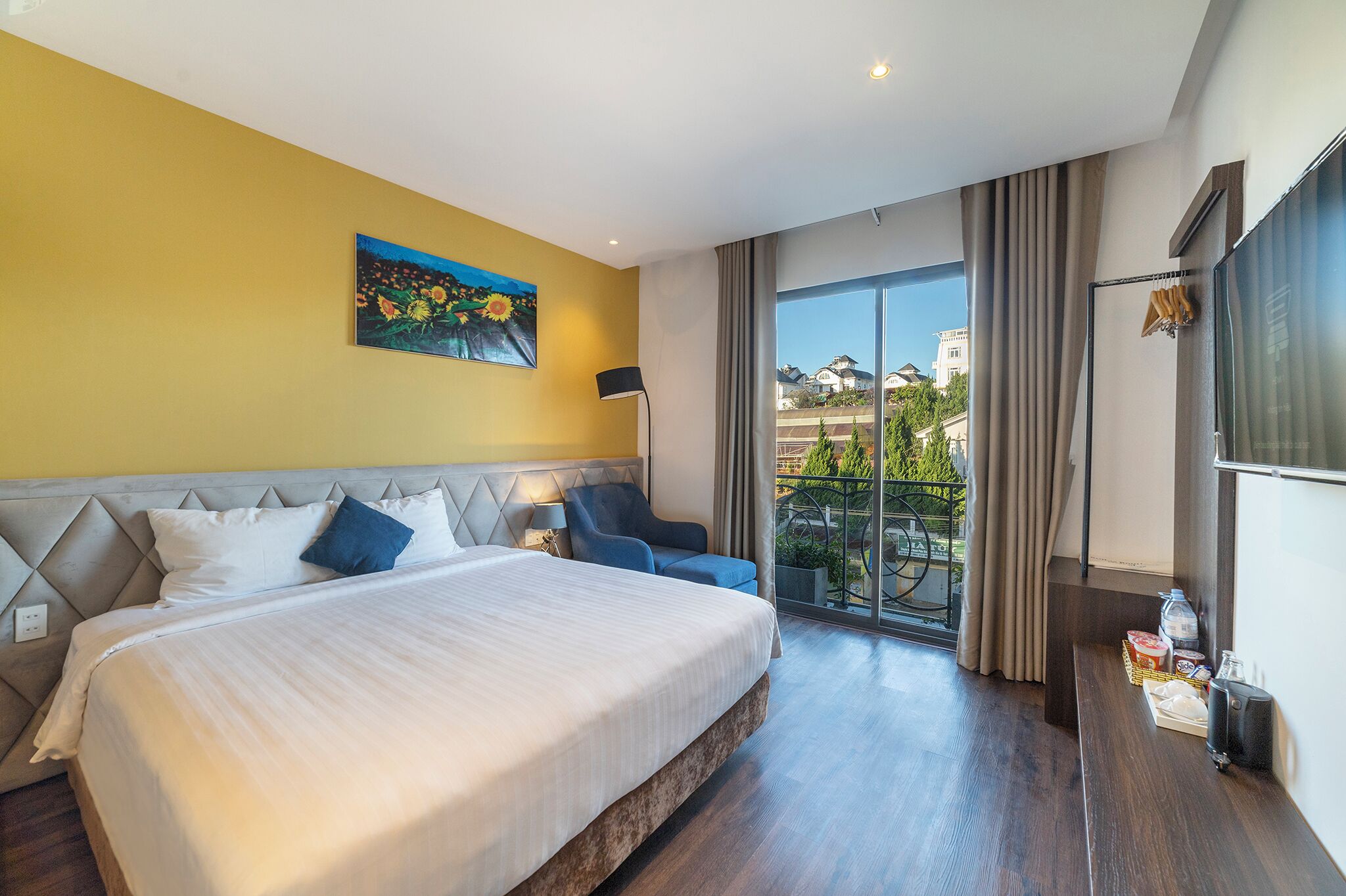 A hotel room with a bed, a chair, a painting, a balcony with curtains, and a TV.
