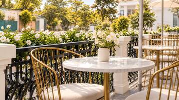 A terrace with round tables and chairs, a white vase with flowers, and a black wrought iron railing.