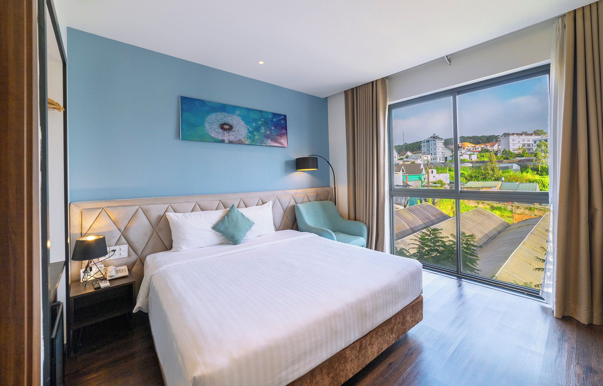 A hotel room with a bed, a nightstand, a lamp, a chair, a painting, and a large window with a view of buildings.