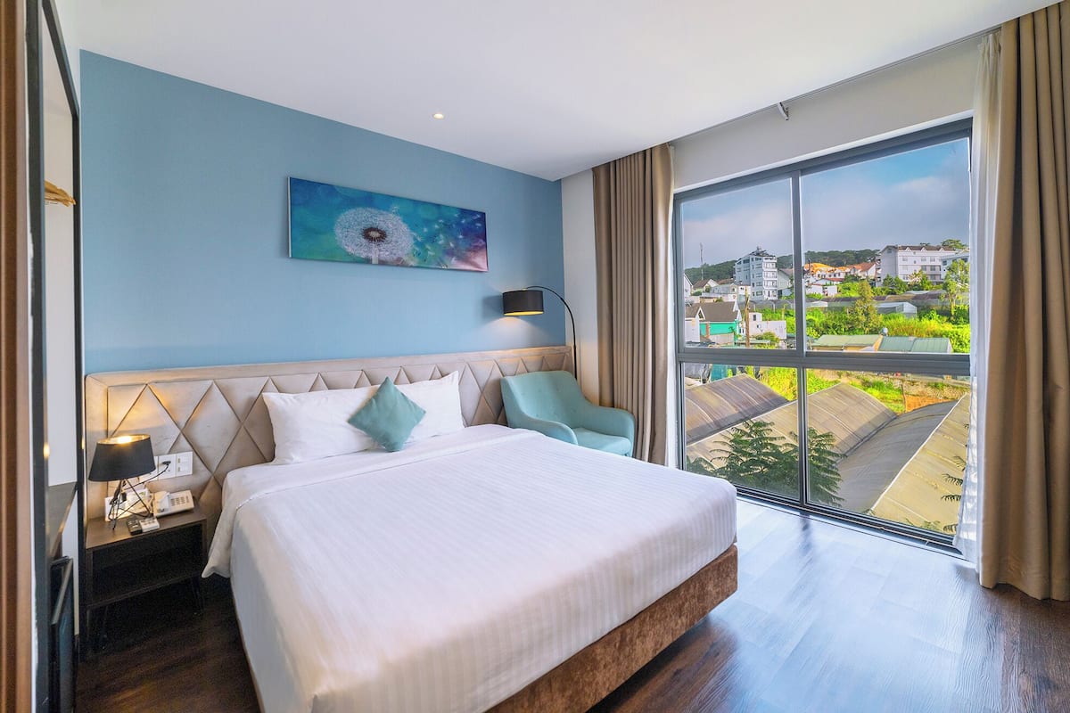 A hotel room with a bed, a nightstand, a lamp, a chair, a painting, and a large window with a view of buildings.