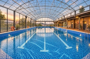 An indoor swimming pool with a glass ceiling, clear poolside area, and a view of the cityscape.