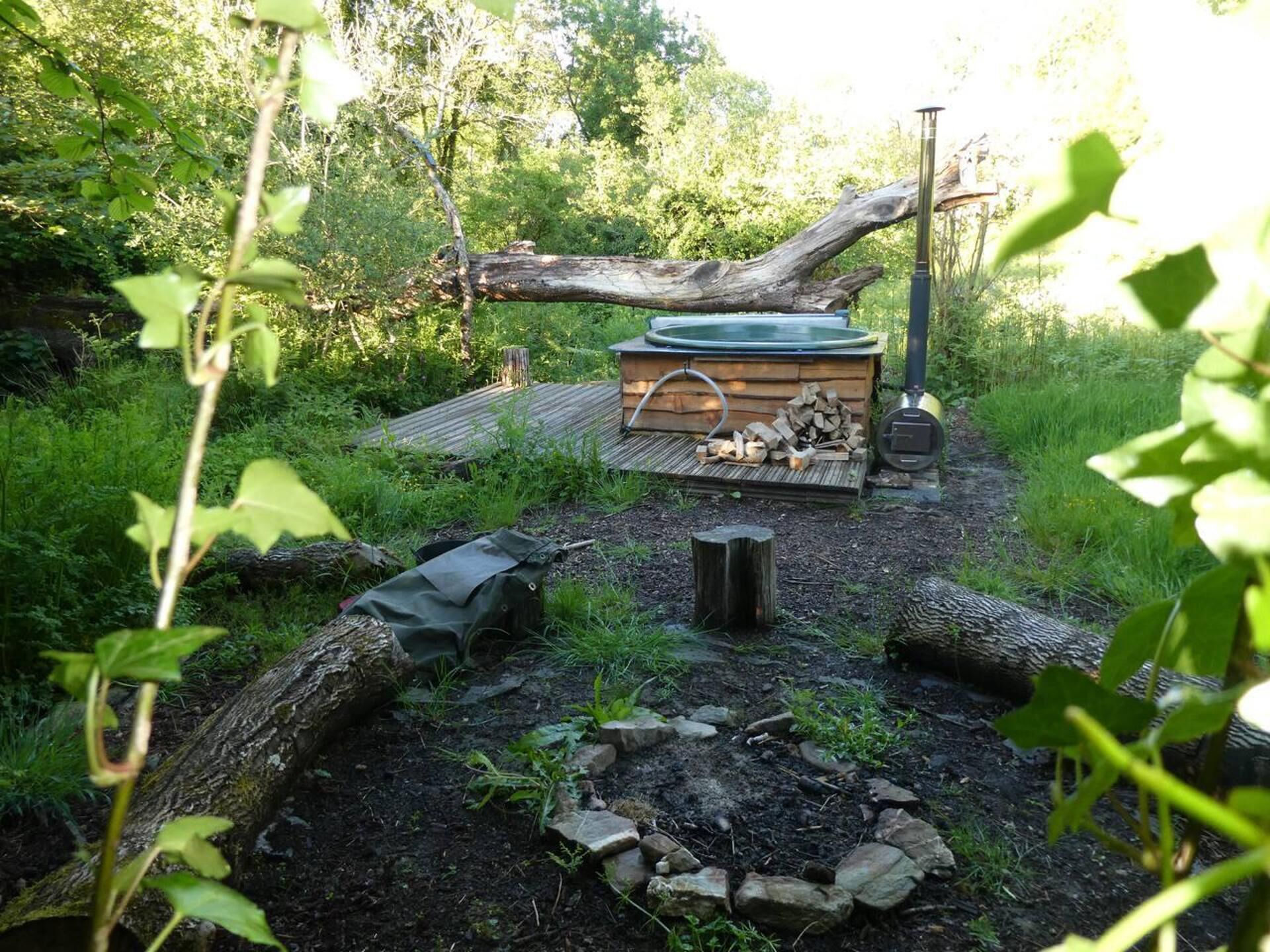 Montaje rústico de jacuzzi al aire libre con terraza de madera, fogón y zona para leña, todo en un entorno boscoso.