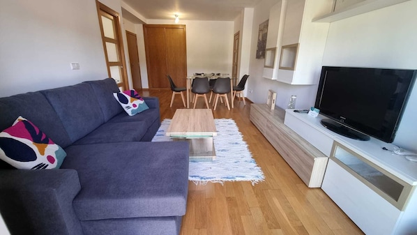 Een moderne woonkamer met een grijze bank, een salontafel, een televisie en een eethoek met stoelen en een tafel.