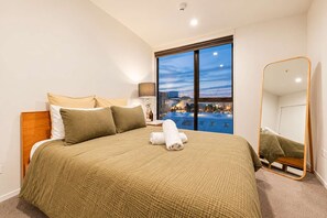 Ein Hotelzimmer mit einem großen Bett, einem Nachttisch, einer Lampe, einem Fenster mit Stadtblick und einem Spiegel.