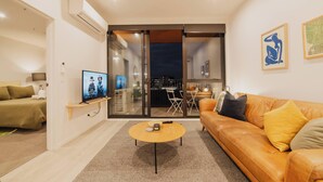 Ein modernes Wohnzimmer mit einer Couch, einem Couchtisch und einem Fernseher. Eine Schiebetür aus Glas führt auf einen Balkon mit Blick auf die Stadt.