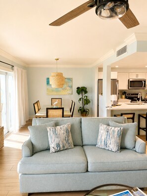 A modern living room with a light blue sofa, a ceiling fan, a dining area with a table and chairs, and a kitchen with white cabinets and appliances.