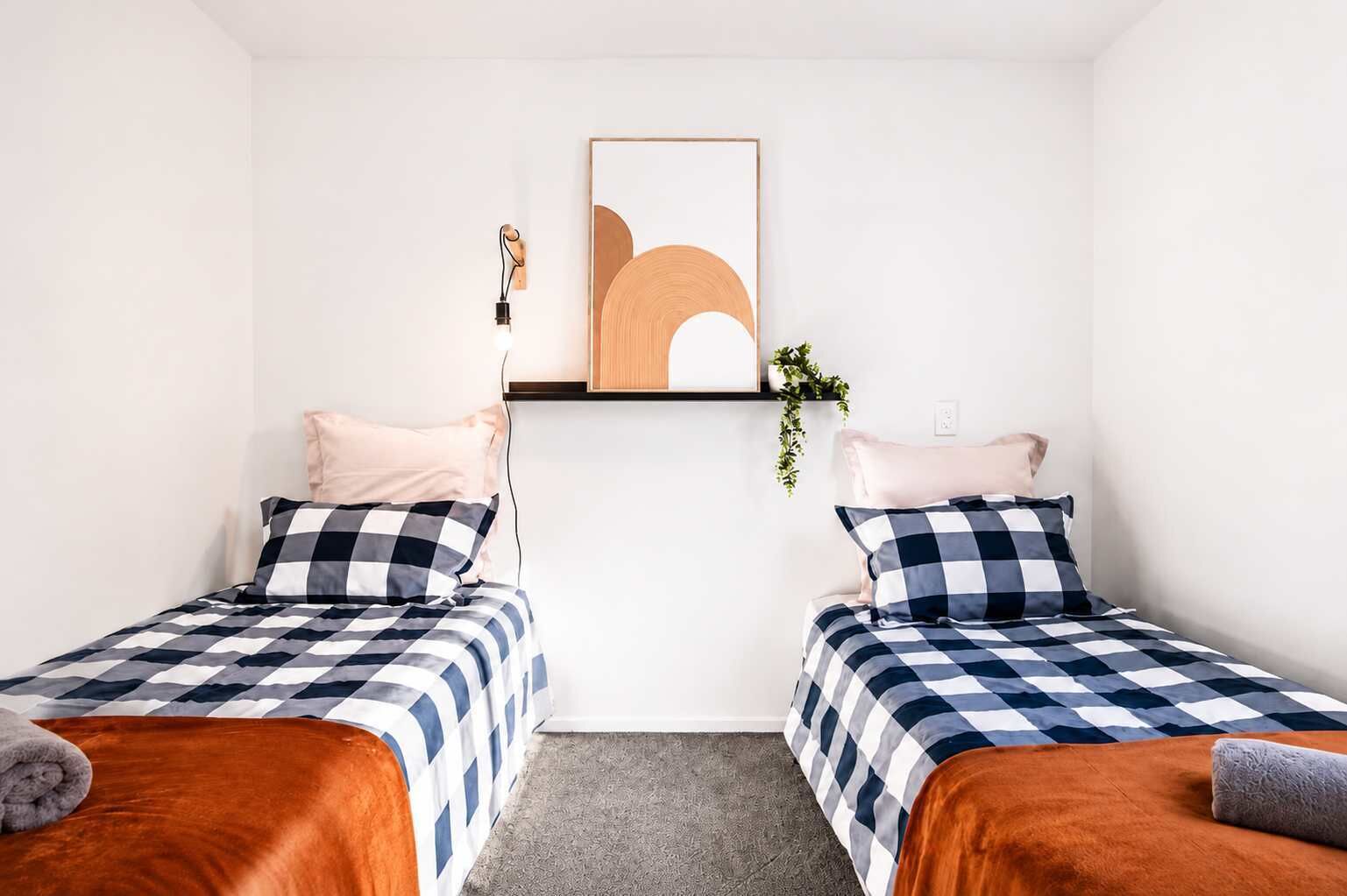 Quarto com duas camas, cada uma com colcha xadrez, uma pequena planta na parede e um luminário acima.