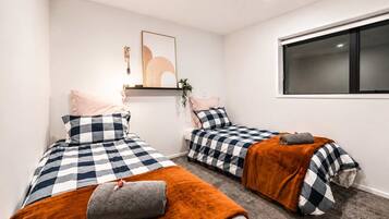 A room with two beds, each with a checkered bedspread and pillows, a window, and a wall-mounted shelf with a plant.