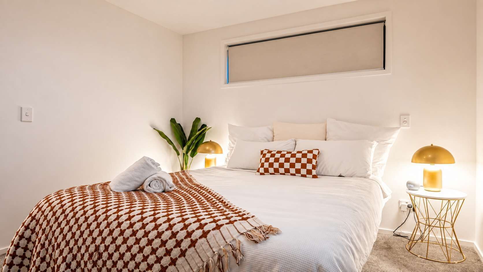 Ein Hotelzimmer mit einem Bett, einer karierten Tagesdecke, einer Pflanze, zwei Nachttischlampen und einer Wandleuchte.