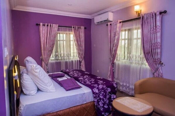 A room with a bed, a chair, and a small table. There are two windows with curtains. - Pearl Hilton Int'l Hotel Limited  (Benin City)