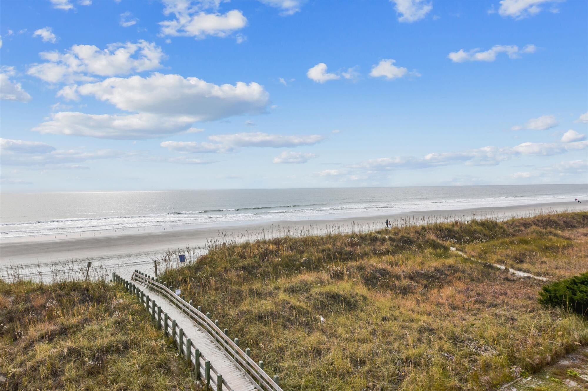 Un paysage côtier avec une plage de sable, des dunes herbeuses et une promenade en bois menant à la rive.