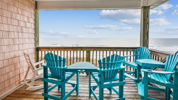 A wooden deck with turquoise Adirondack chairs and a table, overlooking a beach and ocean.