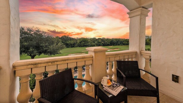 A balcony with a view of a golf course at sunset, featuring two wicker chairs and a table with a cup and glasses. - Spacious home w/ themed rooms, pool & game room (Kissimmee)
