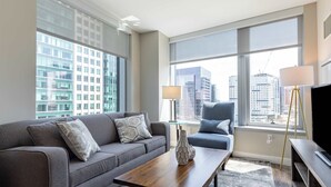 A modern hotel room with a grey sofa, a wooden coffee table, a blue armchair, and a large window offering a city view.