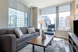 A modern hotel room with a grey sofa, a wooden coffee table, a blue armchair, and a large window offering a city view.