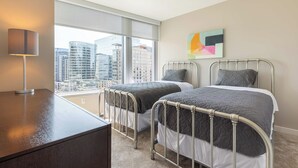 A hotel room with two beds, a desk, a lamp, a large window with a city view, and a colorful abstract painting on the wall.