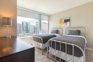 A hotel room with two beds, a desk, a lamp, a large window with a city view, and a colorful abstract painting on the wall.