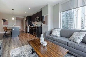 A modern living room with a gray sofa, a wooden coffee table, and a kitchen area with dark cabinets and stainless steel appliances.