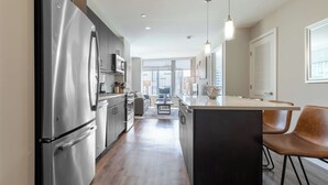 A modern kitchen with a stainless steel refrigerator, wooden flooring, and a central island with chairs.
