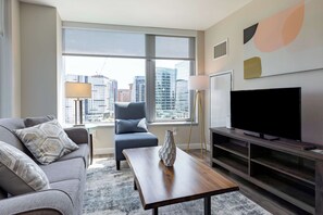 A modern living room with a sofa, a chair, a coffee table, a TV, and a large window offering a city view.