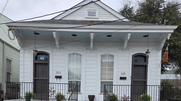 Une maison blanche à deux étages, dotée d’un porche à l’avant, portant les numéros 323 et 325, et un petit jardin agrémenté de plantes en pot.