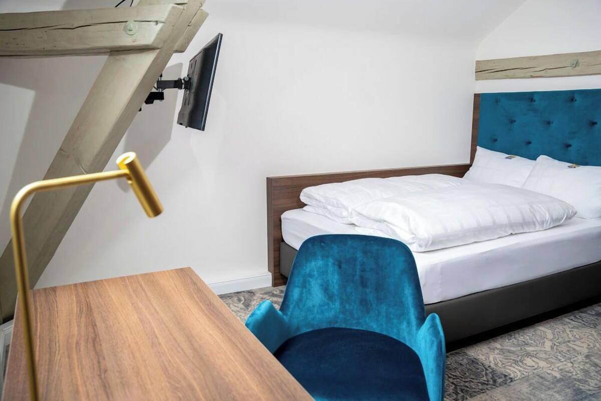 A hotel room with a wooden desk, a blue chair, a bed with white bedding, and a television mounted on the wall.