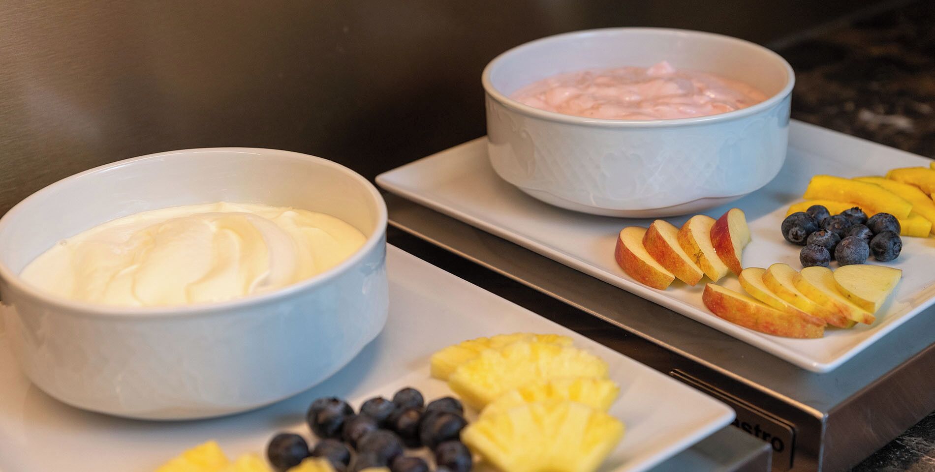 Dos tazones de yogur sobre una bandeja con rodajas de manzana, arándanos y piña.