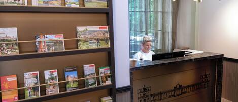 Een receptiebalie met uitzicht op een stadsgezicht door grote ramen, een boekenkast met reisboeken en een moderne kroonluchter.