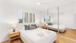 A bedroom with a large bed, bedside tables, a mirror, and a window with shutters. - Unit 8 Charlton on the Esplanade (Torquay)