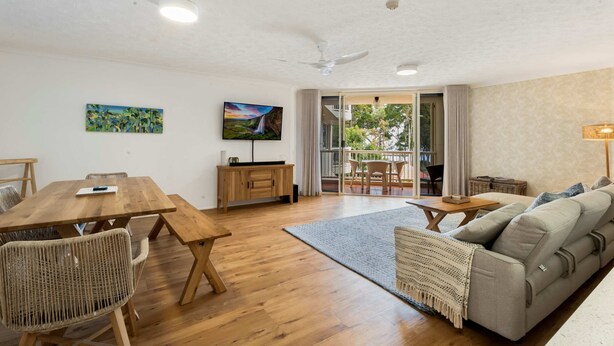 A spacious living area with a wooden dining table, wicker chairs, a sofa, and a television on the wall. - Unit 8 Charlton on the Esplanade (Torquay)