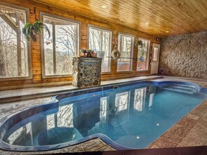 An indoor swimming pool with stone walls and wooden ceiling, large windows, and a stone pillar with a plant. - Unique Peaceful- Romantic, private pool, fireplace, AC-WIFI, delightful Smokies! (Sevierville)