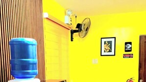 Een kamer met een houten tafel, een waterdispenser, een magnetron, een kleine koelkast, een witte stoel, een gele muur, een ingelijst schilderij, een Wi-Fi sign en een brandblusser.