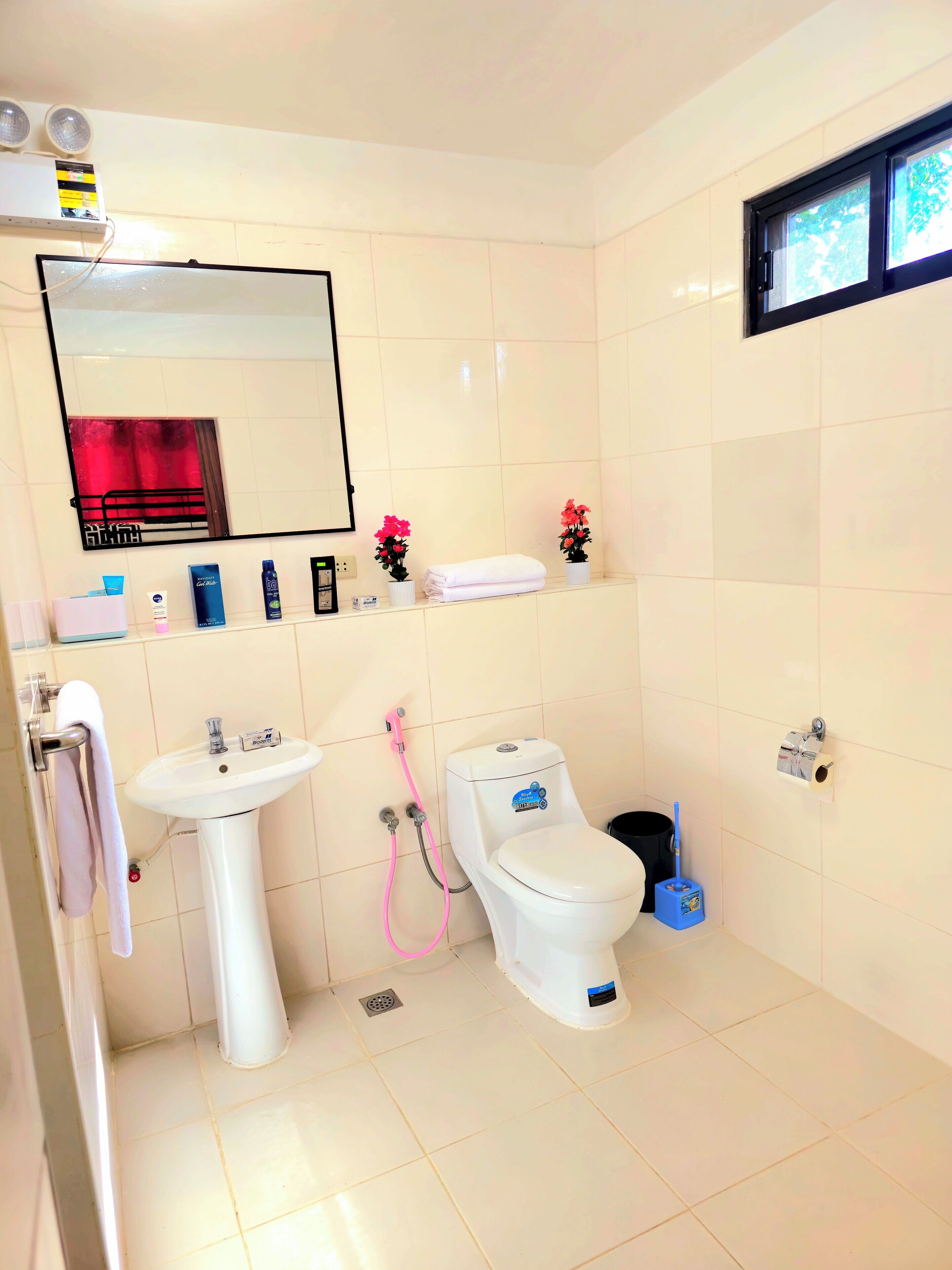 A bathroom with a sink, toilet, and mirror. There are two pink flower pots on the shelf, a white towel hanging on the wall, and a blue bucket on the floor.