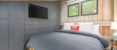 A bedroom with a bed, a TV mounted on the wall, a window with a view of trees, and a wooden ceiling.