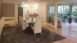 Una cocina moderna con armarios blancos, una zona de comedor con mesa de vidrio y sillas blancas, y un amplio ventanal corredero que da a un balcón con vistas a la vegetación.