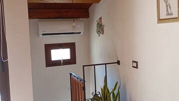 A room with a wooden ceiling, a window, a wall-mounted air conditioner, a framed picture, and a potted plant.